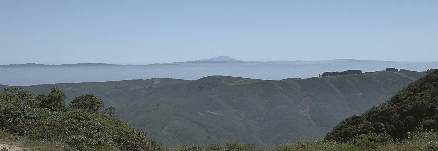 The fog sea on the horizon beyond rolling coastal ridges, the distant peak in full view